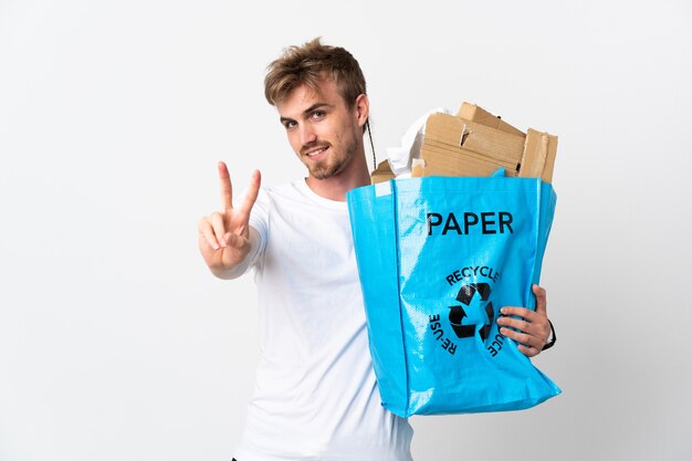 Jonge blonde man met een recycling zak vol papier om te recyclen geïsoleerd op een witte achtergrond glimlachend en overwinningsteken tonen