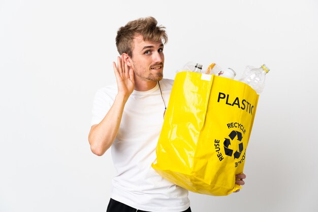 Jonge blonde man met een recycling zak vol papier om te recyclen geïsoleerd op wit luisteren naar iets door hand op het oor te leggen