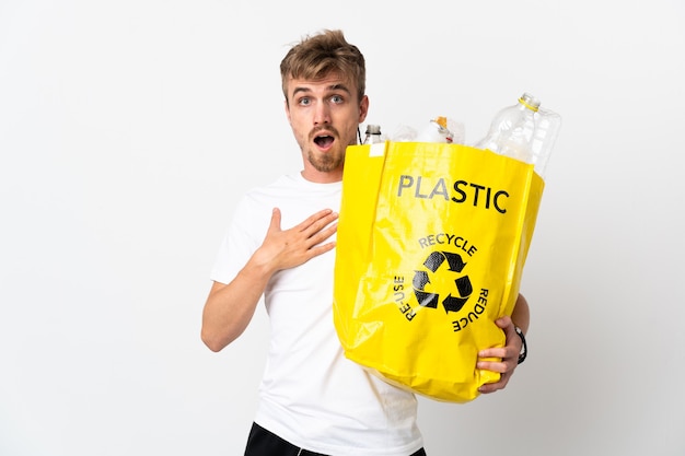 Jonge blonde man met een recycling zak vol papier om te recyclen geïsoleerd op wit verrast en geschokt terwijl hij naar rechts kijkt