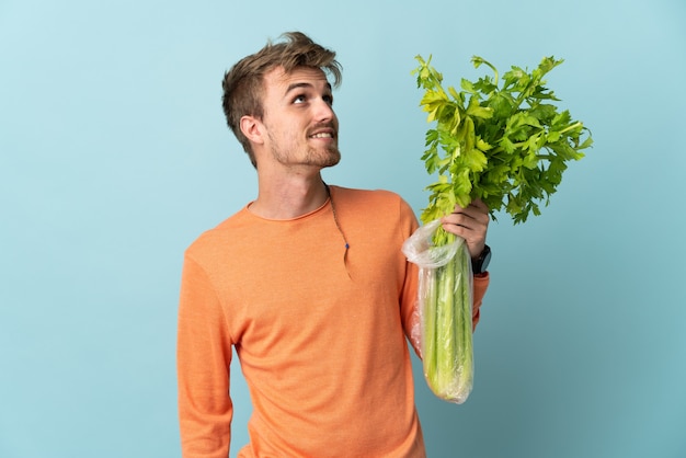 Jonge blonde man met een selderij geïsoleerd op blauwe achtergrond opzoeken tijdens het glimlachen