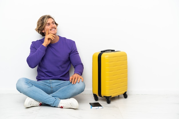 Jonge blonde man met koffer zittend op de vloer een idee te denken tijdens het opzoeken