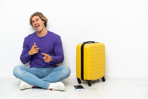 Jonge blonde man met koffer zittend op de vloer naar voren wijzend en lachend