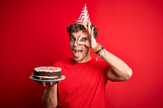 Jonge blonde man met krullend haar met een verjaardagshoed die cake vasthoudt op rode achtergrond met een blij gezicht lachend ok teken doen met de hand op het oog kijkend door de vingers