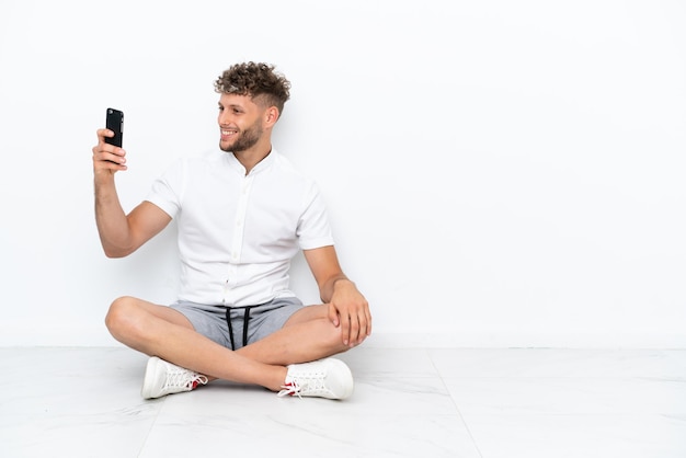 Jonge blonde man zittend op de vloer geïsoleerd op een witte achtergrond maken een selfie