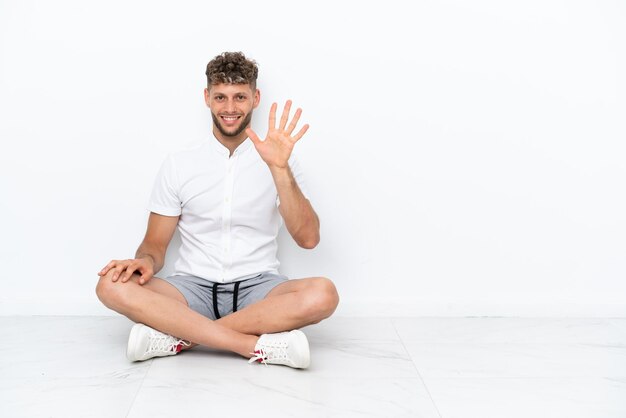 Jonge blonde man zittend op de vloer geïsoleerd op een witte achtergrond telt vijf met vingers