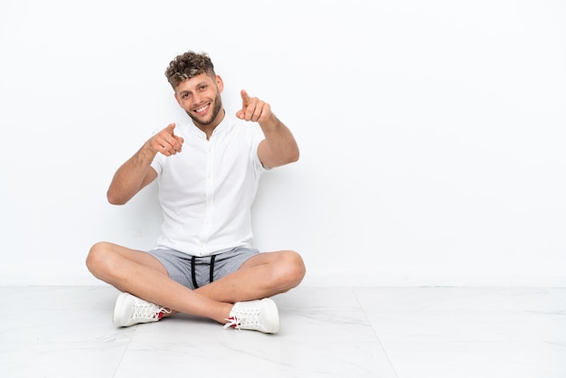 Jonge blonde man zittend op de vloer geïsoleerd op een witte achtergrond wijst vinger naar je terwijl hij lacht