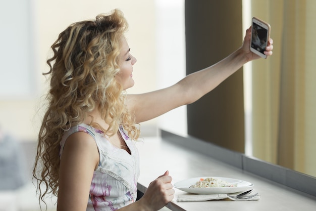 Jonge blonde meid die zelfportret maakt op haar smartphone digitale camera terwijl ze tijdens de lunchpauze in café zit.