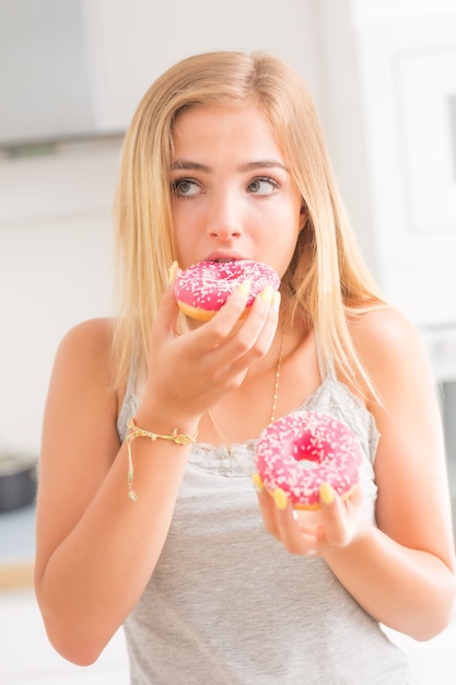 Jonge blonde meid eet roze donuts in de keuken van het huis met smaakemoties.