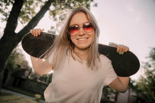 Foto jonge blonde meid houdt haar skateboard vast, kijkt naar de camera en glimlacht.