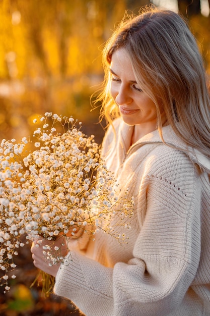 Jonge blonde meid met bloemen die in het herfstpark in de buurt van het meer staat Vrouw draagt beige trui Meisje poseert voor een foto op zonnige dag