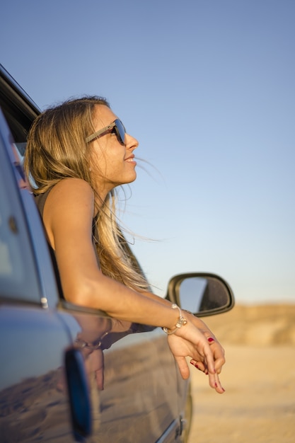 Jonge blonde meid met een zonnebril kijkt uit de auto in een woestijn in Las Bardenas Reales, Spanje