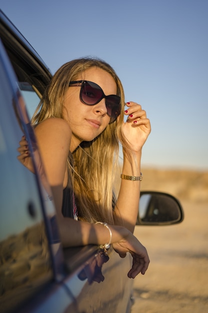 Jonge blonde meid met een zonnebril kijkt uit de auto in een woestijn in Las Bardenas Reales, Spanje