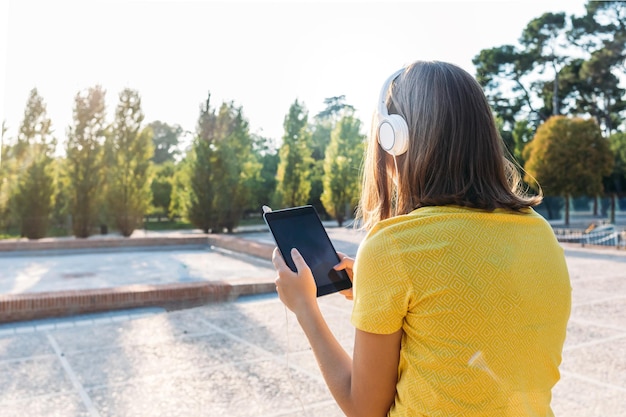 Jonge blonde meid met haar rug naar haar toe, geel t-shirt en witte koptelefoon dragend, glimlachend en kijkend naar haar tablet, zittend op een reling. Technologie, lifestyle en ontspanningsconcept