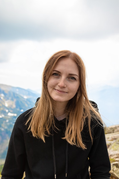 Jonge blonde meisjesreiziger met een blauwe rugzak die en van het groene berglandschap wordt opgeheven geniet