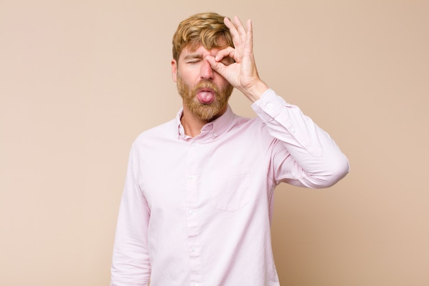 Jonge blonde mens die gelukkig met grappig gezicht glimlacht, een grapje maakt en door kijkgaatje kijkt, dat geheimen tegen vlakke muur bespioneert