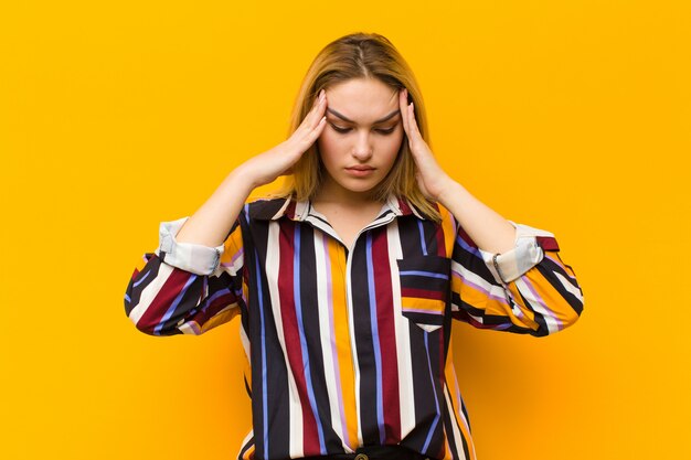 Jonge blonde mooie vrouw die beklemtoond en gefrustreerd kijkt, werkend onder druk met hoofdpijn en verontrust met problemen tegen oranje muur