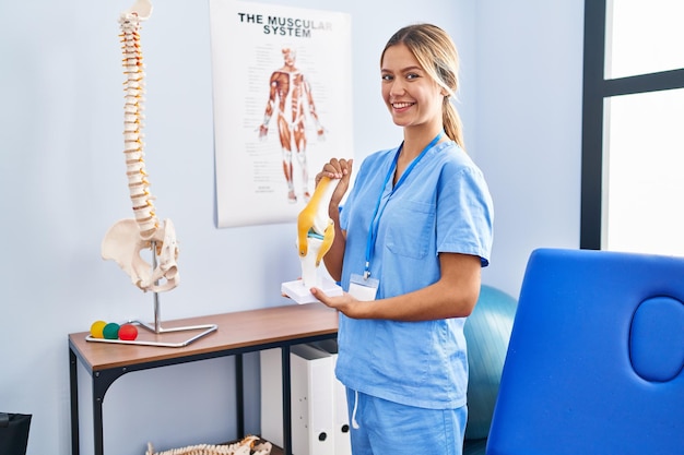 Jonge blonde orthopedische vrouw met anatomisch model van kniegewricht glimlachend met een gelukkige en koele glimlach op het gezicht met tanden