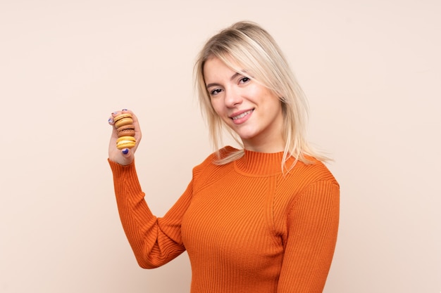 Jonge blonde Russische vrouw over geïsoleerde muur die kleurrijke Franse macarons houdt en veel glimlacht