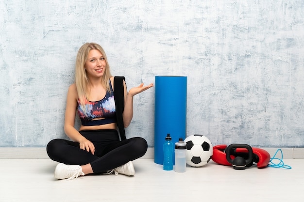 Jonge blonde sport vrouw zittend op de vloer uitbreiding van de handen aan de zijkant voor het uitnodigen om te komen