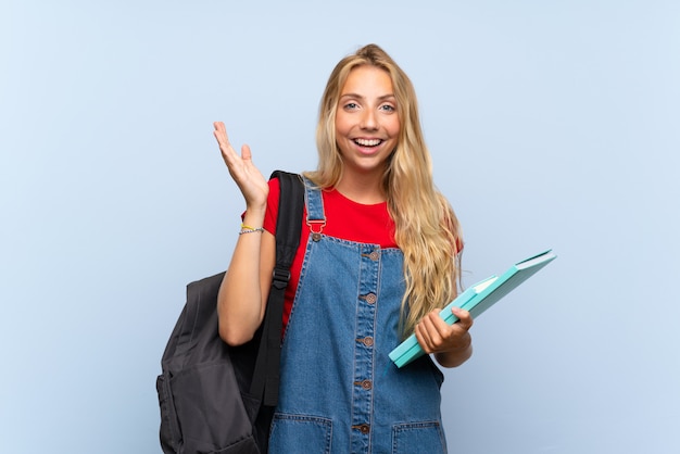 Jonge blonde studentenvrouw over geïsoleerde blauwe muur met verrassingsgelaatsuitdrukking