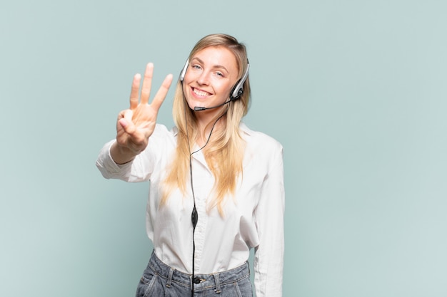 Jonge blonde telemarketeervrouw die lacht en er vriendelijk uitziet, nummer drie of derde toont met de hand naar voren, aftellend