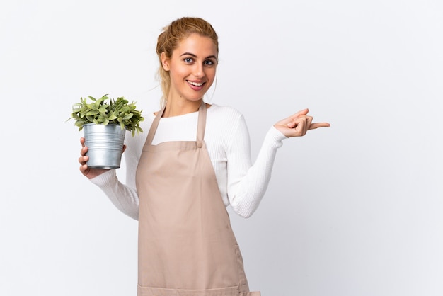 Jonge blonde tuinman vrouw meisje met een plant over geïsoleerde witte muur wijzende vinger naar de zijkant