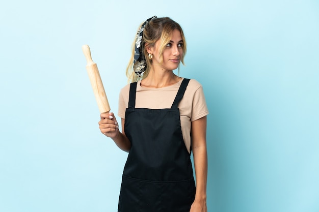 Jonge blonde Uruguayaanse kokende vrouw die op blauwe muur wordt geïsoleerd die aan de kant kijkt