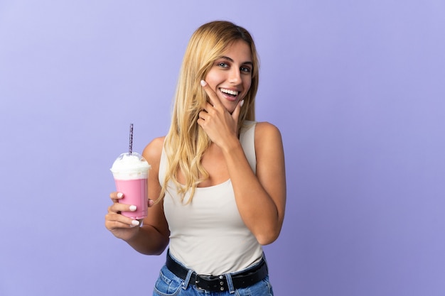 Jonge blonde Uruguayaanse vrouw met aardbeimilkshake geïsoleerd op paarse muur blij en glimlachend