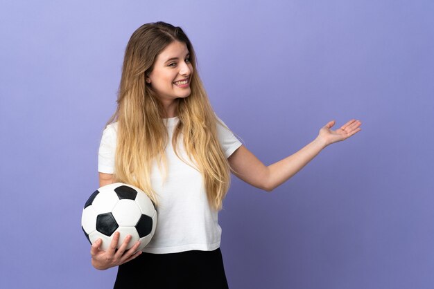 Jonge blonde voetballervrouw die op purpere muur wordt geïsoleerd die handen naar de kant uitbreidt om uit te nodigen om te komen