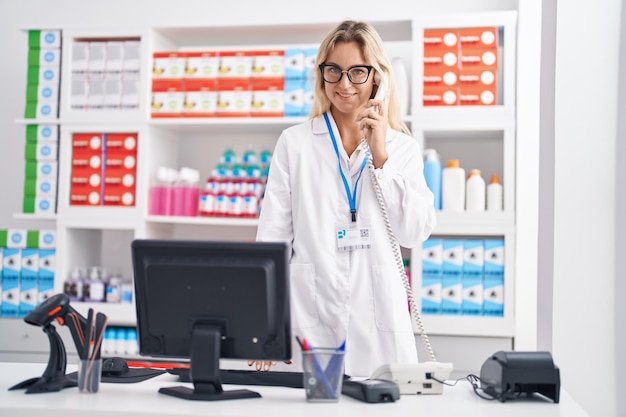 Jonge blonde vrouw apotheker praten aan de telefoon met behulp van computer bij apotheek