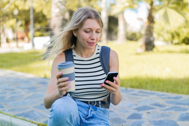 Jonge blonde vrouw buitenshuis met behulp van mobiele telefoon en het houden van een afhaalkoffie