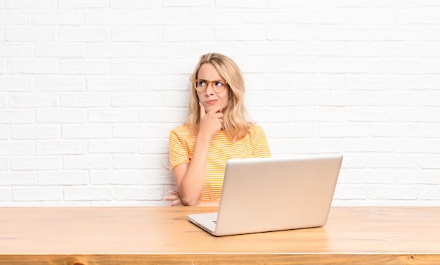 Jonge blonde vrouw denken, twijfelachtig en verward voelen, met verschillende opties, af welke beslissing te nemen met behulp van een laptop