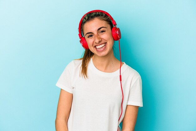 Jonge blonde vrouw die aan muziek op hoofdtelefoons luistert die op blauwe muur wordt geïsoleerd gelukkig, glimlachend en vrolijk