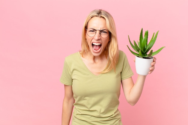 Jonge blonde vrouw die agressief schreeuwt, erg boos kijkt en een cactus vasthoudt