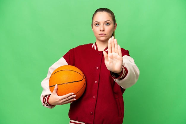 Jonge blonde vrouw die basketbal speelt over geïsoleerde chroma key achtergrond die stopgebaar maakt