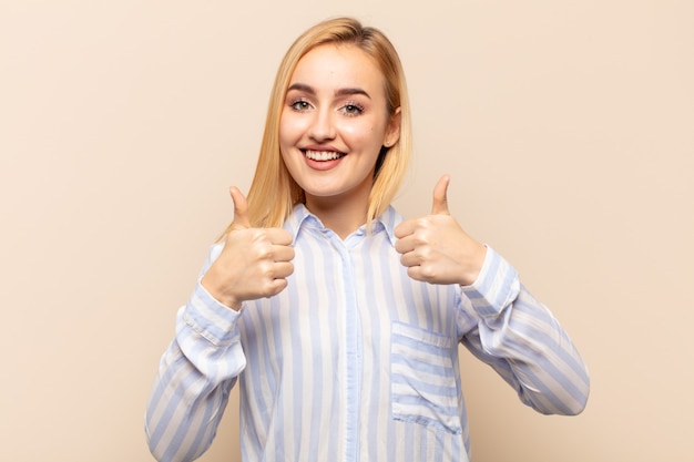 Jonge blonde vrouw die breed kijkt gelukkig, positief, zelfverzekerd en succesvol, met beide duimen omhoog