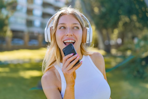 Jonge blonde vrouw die buiten muziek luistert met een mobiel en zingt