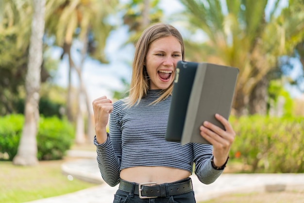 Jonge blonde vrouw die buitenshuis een tablet vasthoudt en een overwinning viert