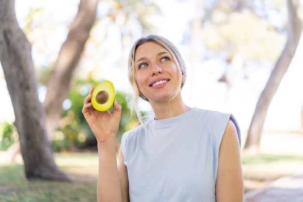 Foto jonge blonde vrouw die een avocado vasthoudt terwijl ze glimlacht