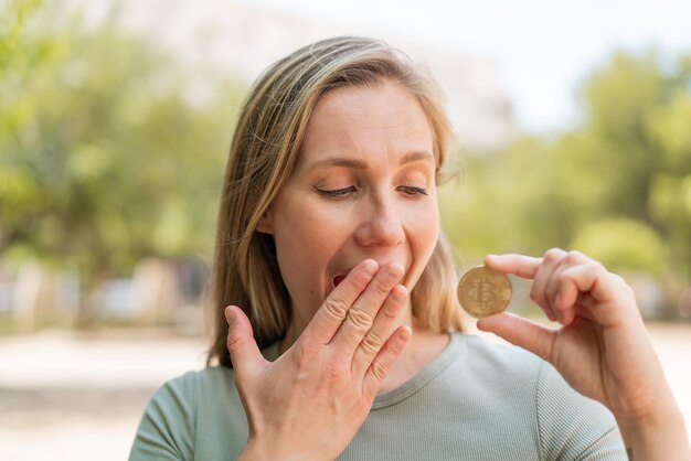 Foto jonge blonde vrouw die een bitcoin buiten vasthoudt met verbazing en geschokte gezichtsuitdrukking