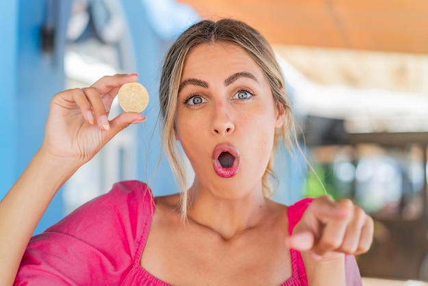 Foto jonge blonde vrouw die een bitcoin in de buitenlucht houdt, verrast en wijst naar voren