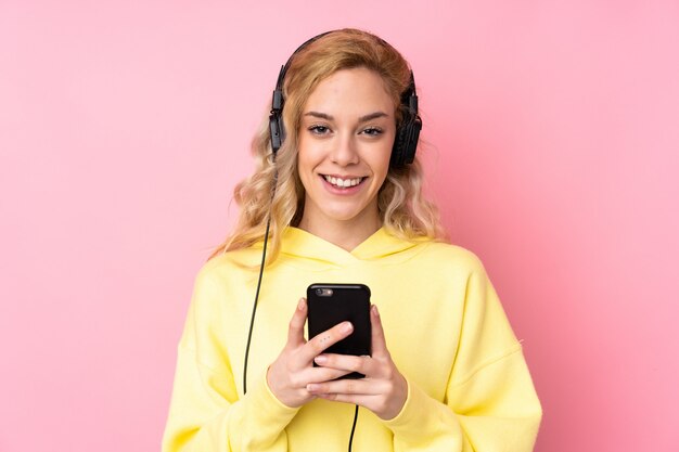 Jonge blonde vrouw die een sweatshirt op roze het luisteren muziek met mobiel draagt en voorzijde kijkt