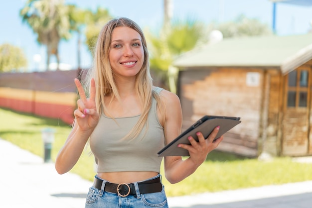 Jonge blonde vrouw die een tablet vasthoudt in de open lucht glimlachend en overwinningsteken tonend