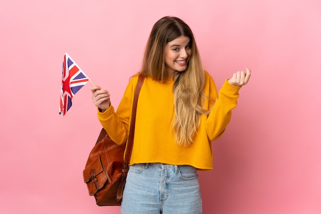 Jonge blonde vrouw die een vlag van het Verenigd Koninkrijk houdt die op witte muur wordt geïsoleerd die geldgebaar maakt