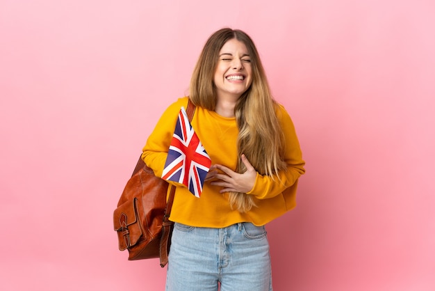 Jonge blonde vrouw die een vlag van het Verenigd Koninkrijk houdt die op witte ruimte wordt geïsoleerd die veel glimlacht