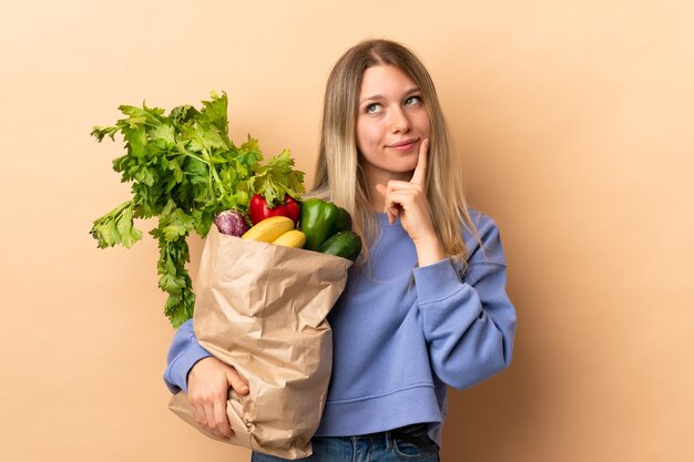 Jonge blonde vrouw die een zak vol groenten over geïsoleerde muur houdt die een idee denken