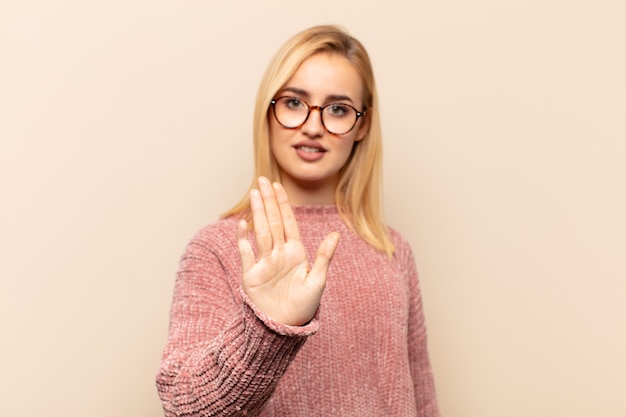 Jonge blonde vrouw die ernstig, streng, ontevreden en boos kijkt die open palm toont die stopgebaar maakt