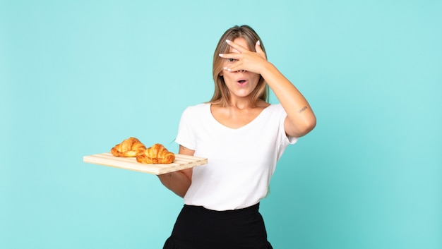 Jonge blonde vrouw die geschokt, bang of doodsbang kijkt, haar gezicht bedekt met de hand en een croissantblad vasthoudt