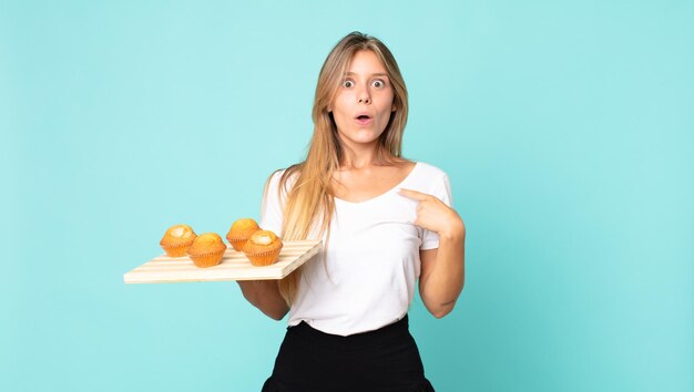 Jonge blonde vrouw die geschokt en verrast kijkt met wijd open mond wijzend naar zichzelf en een muffins troy vasthoudt