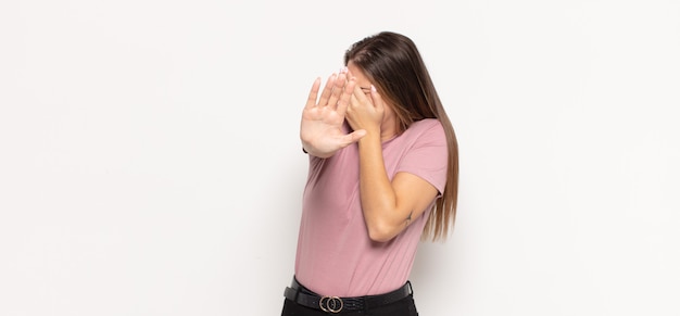 Jonge blonde vrouw die gezicht bedekt met hand en andere hand naar voren zet om te stoppen, foto's of afbeeldingen weigeren